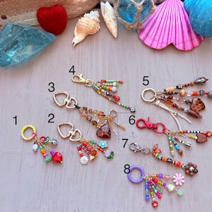 May include: A selection of beaded keyrings with diverse designs. The keyrings feature colourful beads, charms, and metal hardware. Some have heart-shaped charms, while others have flower or tassel accents. The keyrings are arranged on a light-coloured surface.