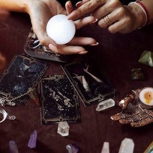 May include: A white crystal ball is held between two hands, surrounded by tarot cards, crystals, and a candle. The scene is set on a dark wooden surface, creating a mystical and spiritual atmosphere.