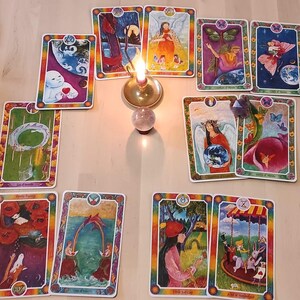 Op de afbeelding: Een tarotkaart lay-out met kleurrijke kaarten met illustraties van engelen, figuren en symbolische beelden. Een brandende kaars en een kristallen bol staan centraal, wat een warme gloed creëert.
