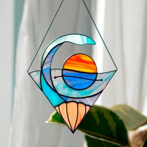 Peut inclure: Un capteur de soleil en vitrail représentant une vague et un coucher de soleil. Le design utilise des teintes de bleu, d'orange et de jaune, encadré dans un losange. Le capteur de soleil est suspendu par une chaîne.