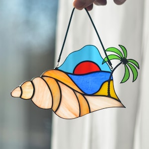 Peut inclure: Un attrape-soleil en verre teinté en forme de coquille avec un motif de coucher de soleil, d'océan et de palmier. La coquille est orange et blanche, le coucher de soleil est rouge et orange, l'océan est bleu et le palmier est vert.