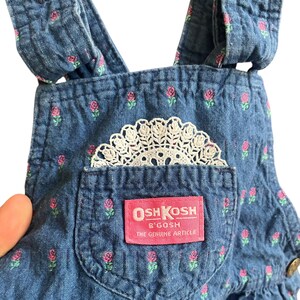 Op de afbeelding: Close-up van een spijkerbroek met een bloemenpatroon van roze rozen en groene bladeren. Een wit kanten kleedje zit in de zak. Het label luidt "OSH KOSH B'GOSH THE GENUINE ARTICLE."