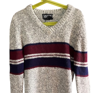 Puede incluir: Un suéter gris jaspeado con cuello en V con un diseño de rayas horizontales en burdeos, blanco y azul marino. El suéter de manga larga tiene puños y dobladillo acanalados. La etiqueta dice "CHRIS TOPHER RAN".