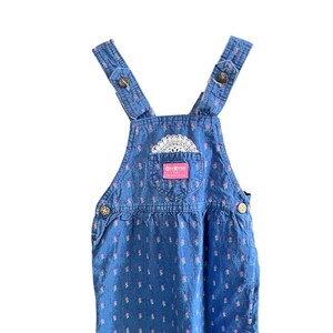 Op de afbeelding: Blauwe denim overall jurk met roze bloemenprint en witte kanten details. Voorzien van verstelbare bandjes met knoopsluitingen en een voorzak met het OshKosh-logo. Een jurk in vintage stijl.