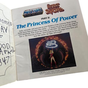Może przedstawiać: Otwarta strona książki z "Masters of the Universe" i "The Princess of Power". Strona zawiera tekst i ilustrację postaci w czerwonym stroju. Książka jest otwarta na białej powierzchni.