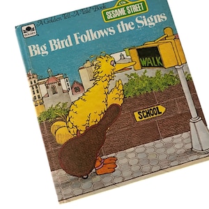 以下が含まれることがあります： 「Big Bird Follows the Signs」のゴールデン・テル・ア・テイル・ブックのヴィンテージ本。表紙には、セサミストリートの黄色いキャラクター、ビッグバードが歩道で横断歩道付近を歩いている様子が描かれています。本は青色の背景にタイトルとセサミストリートのロゴがあります。