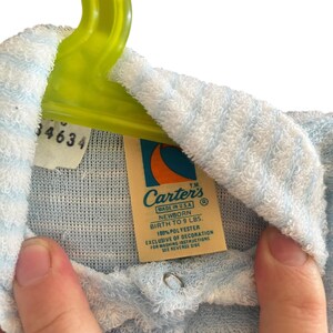 Może przedstawiać: Jasnoniebieski i biały str&oacute;j dla noworodka z metką z napisem "Carter's". Na metce widnieje r&oacute;wnież napis "Urodzenie do 4,1 kg" i "100% poliester". Str&oacute;j wisi na zielonym wieszaku.