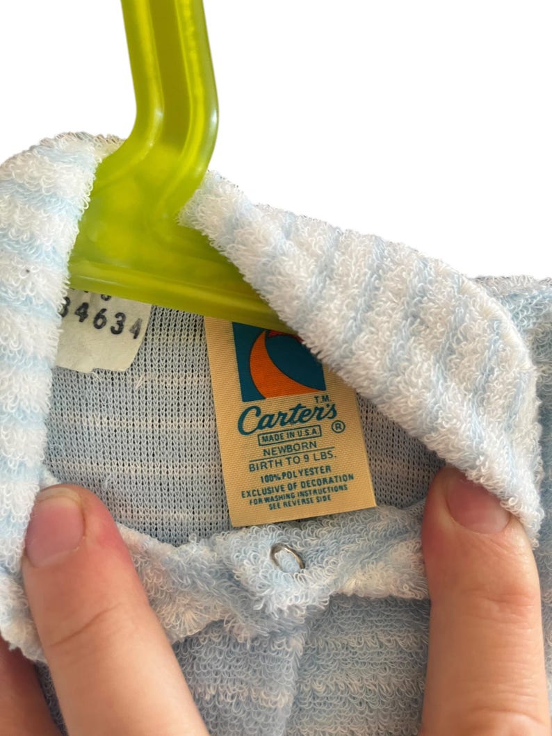 Może przedstawiać: Jasnoniebieski i biały str&oacute;j dla noworodka z metką z napisem "Carter's". Na metce widnieje r&oacute;wnież napis "Urodzenie do 4,1 kg" i "100% poliester". Str&oacute;j wisi na zielonym wieszaku.