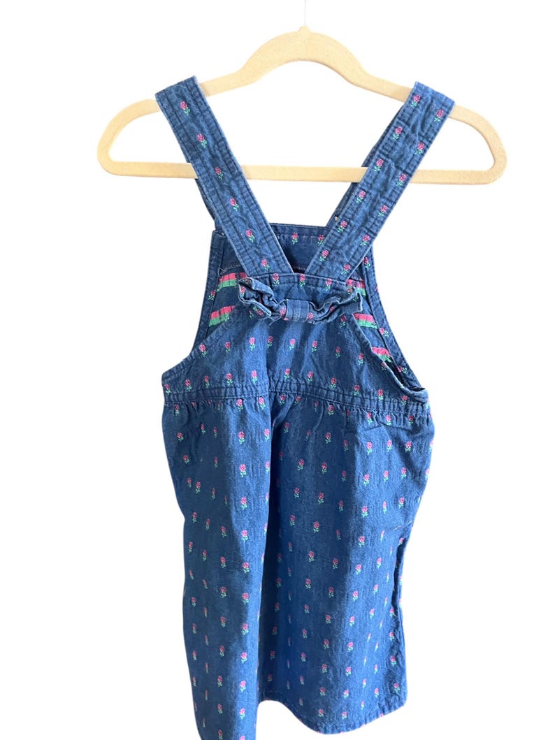 Op de afbeelding: Blauwe denim overall jurk met roze bloemenpatroon. De jurk heeft verstelbare bandjes en een decoratieve strik aan de achterkant. De jurk hangt aan een houten kleerhanger.