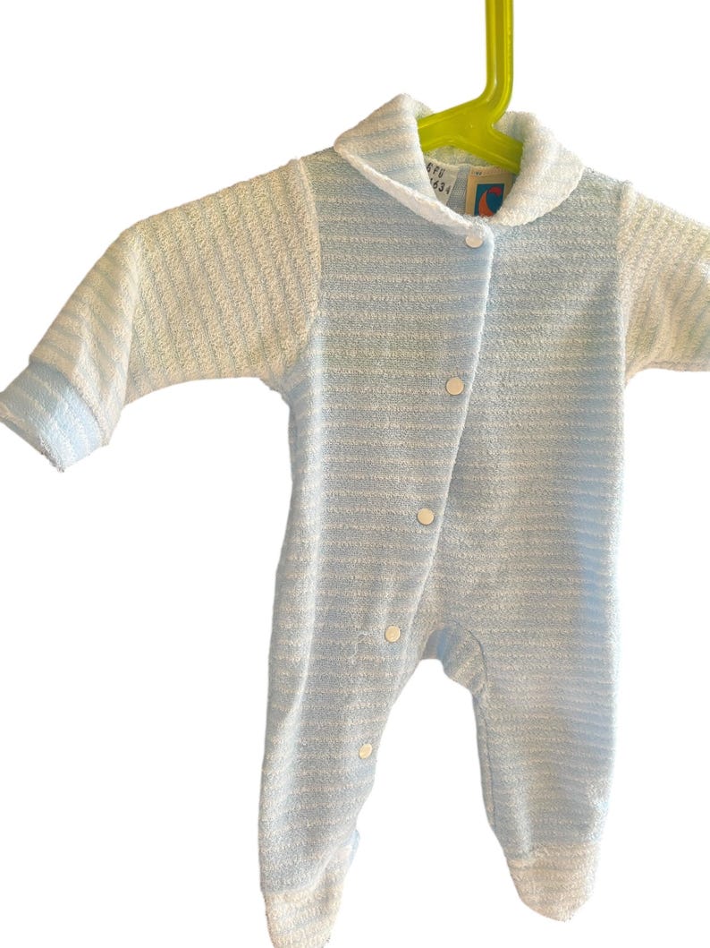 Może przedstawiać: Niemowlęcy pajacyk w niebiesko-białe paski, z kołnierzykiem i długimi rękawami. Pajacyk ma białe guziki i wisi na ż&oacute;łtym wieszaku. Tkanina wydaje się być miękkim frotte. Idealny dla niemowląt.