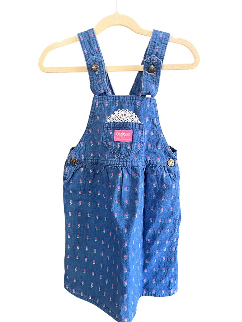 Op de afbeelding: Blauwe denim overall jurk met een bloemenpatroon. De jurk heeft verstelbare bandjes, decoratieve knopen en een zak met het OshKosh-logo. Een wit kanten detail bevindt zich boven de zak. De jurk hangt aan een houten hanger.