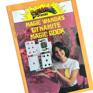 May include: A paperback book titled Magic Wanda's Dynamite Magic Book with an orange border. The cover features a person in a white t-shirt, four playing cards, and the text Dynamite Books and Chip Lovitt against a pink and black background.