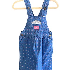 Op de afbeelding: Blauwe denim OshKosh B'gosh overall jurk met een bloemenpatroon. De jurk heeft verstelbare bandjes, knoopsluitingen en een voorzak met een kanten detail. De merknaam is zichtbaar op de zak.
