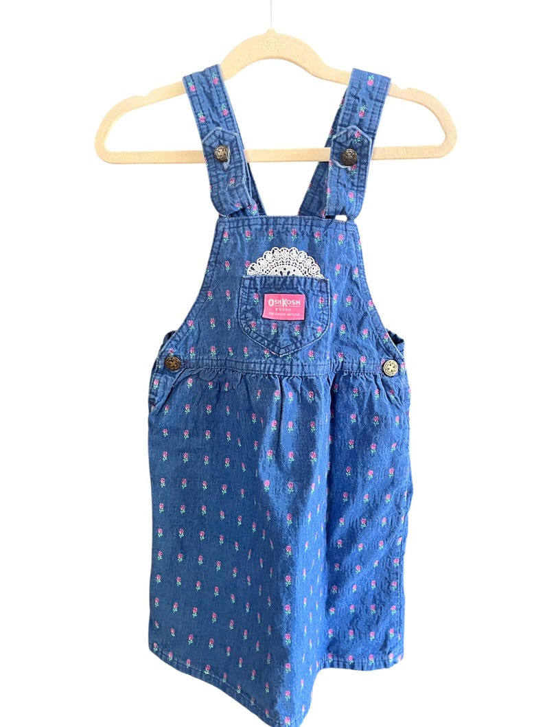 Op de afbeelding: Blauwe denim OshKosh B'gosh overall jurk met een bloemenpatroon. De jurk heeft verstelbare bandjes, knoopsluitingen en een voorzak met een kanten detail. De merknaam is zichtbaar op de zak.