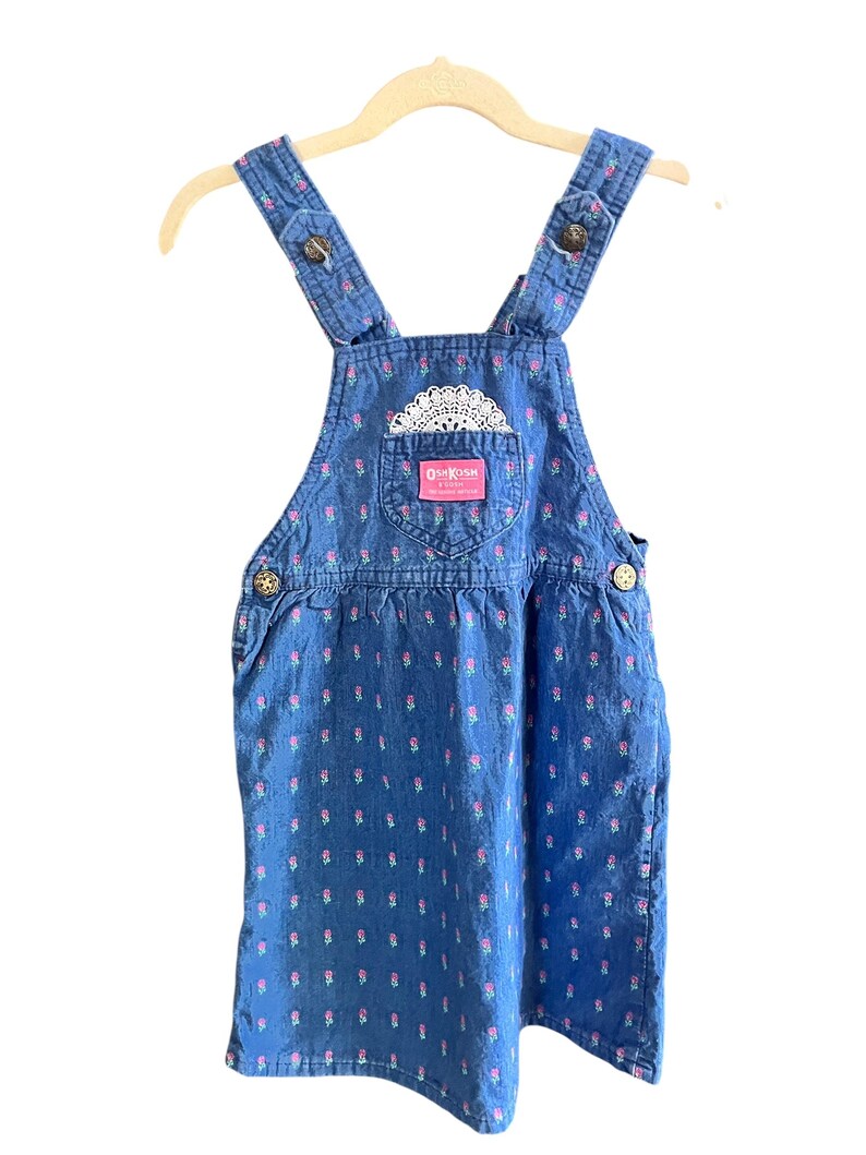 Op de afbeelding: Blauwe denim overall jurk met roze bloemenprint en een wit kanten detail. Voorzien van verstelbare bandjes met knopen en een zak met het OshKosh B'gosh logo. De jurk hangt aan een houten kleerhanger.