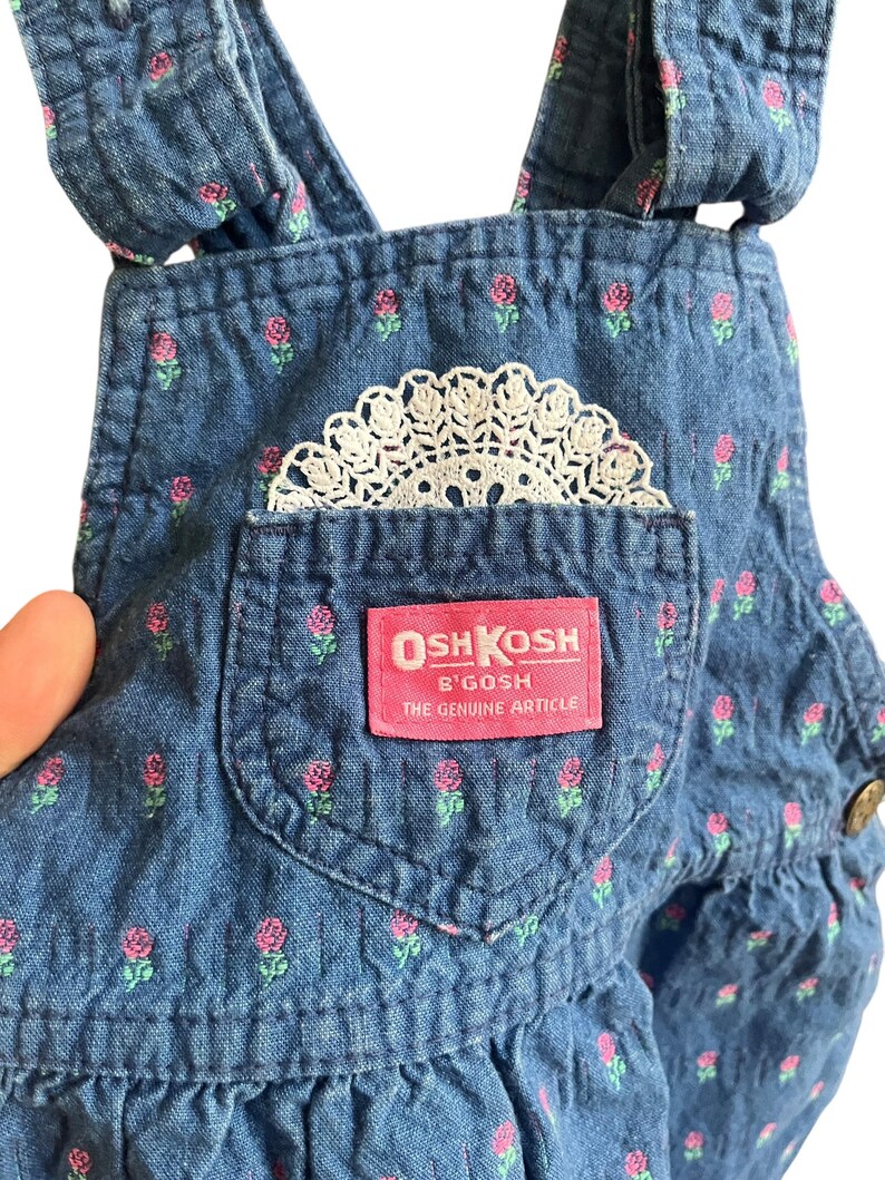 Op de afbeelding: Close-up van een spijkerbroek met een bloemenpatroon van roze rozen en groene bladeren. Een wit kanten kleedje zit in de zak. Het label luidt "OSH KOSH B'GOSH THE GENUINE ARTICLE."