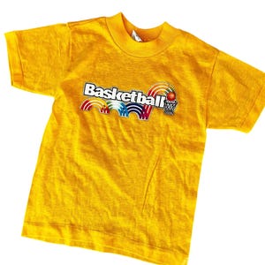 May include: A yellow t-shirt featuring the word "Basketball" in a white, outlined font, with a basketball and net graphic. The design includes rainbow-colored arches. The shirt is short-sleeved and has a crew neck.