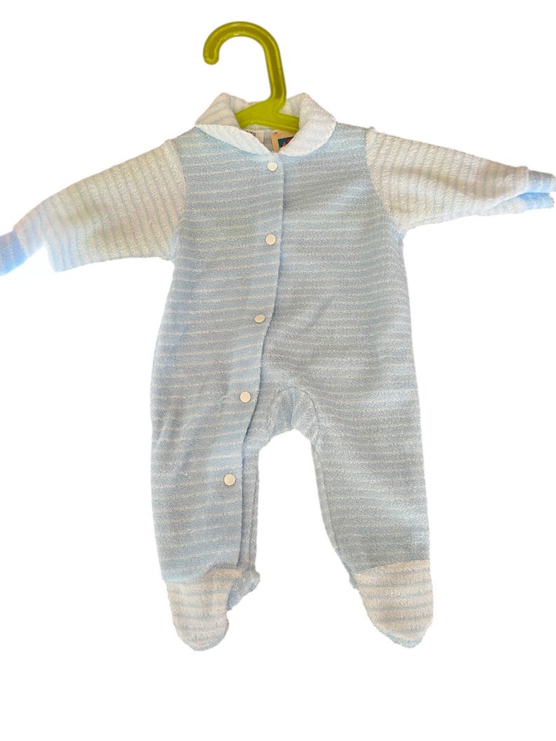 Może przedstawiać: Jasnoniebieski pajacyk dla dziecka z białym kołnierzem i mankietami, zawieszony na zielonym wieszaku. Pajacyk ma zapięcie na guziki z przodu i wz&oacute;r w paski. Materiał wydaje się miękki i teksturowany.