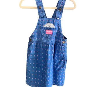 Op de afbeelding: Blauwe denim overall jurk met een bloemenpatroon en een zak met een wit kanten detail. De jurk heeft verstelbare bandjes en knoopsluitingen. Het merklabel "OshKosh" is zichtbaar.