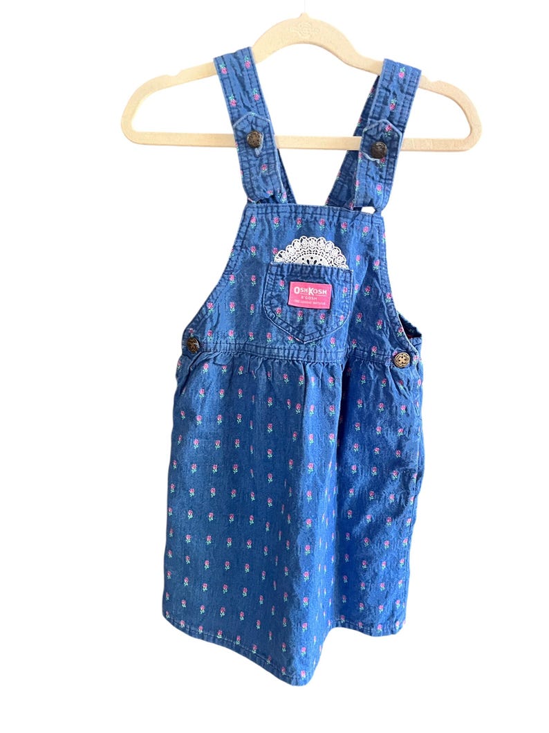 Op de afbeelding: Blauwe denim overall jurk met een bloemenpatroon en een zak met een wit kanten detail. De jurk heeft verstelbare bandjes en knoopsluitingen. Het merklabel "OshKosh" is zichtbaar.