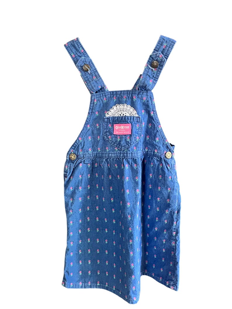 Op de afbeelding: Blauwe denim overall jurk met roze bloemenprint en witte kanten details. Voorzien van verstelbare bandjes met knoopsluitingen en een voorzak met het OshKosh-logo. Een jurk in vintage stijl.