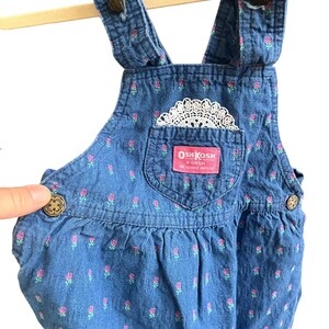 Op de afbeelding: Blauwe denim overall met roze bloemenprint en een wit kanten zakdetail. Voorzien van het label "OshKosh B'Gosh". De overall heeft verstelbare bandjes en knoopsluitingen. Een kledingstuk in vintage stijl.