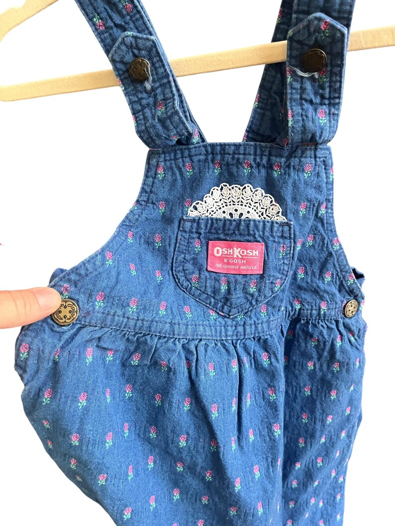 Op de afbeelding: Blauwe denim overall met roze bloemenprint en een wit kanten zakdetail. Voorzien van het label "OshKosh B'Gosh". De overall heeft verstelbare bandjes en knoopsluitingen. Een kledingstuk in vintage stijl.