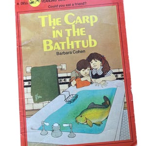 May include: A vintage children's book cover with a red background. The title is "The Carp in the Bathtub" by Barbara Cohen. The cover features an illustration of two children looking into a bathtub with a large carp swimming inside.