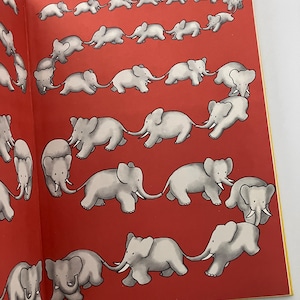 May include: A red book with a yellow border, featuring a series of illustrations of elephants walking in a line, with each elephant holding the tail of the elephant in front of it. The elephants are all facing the same direction and are drawn in a simple, cartoon style.