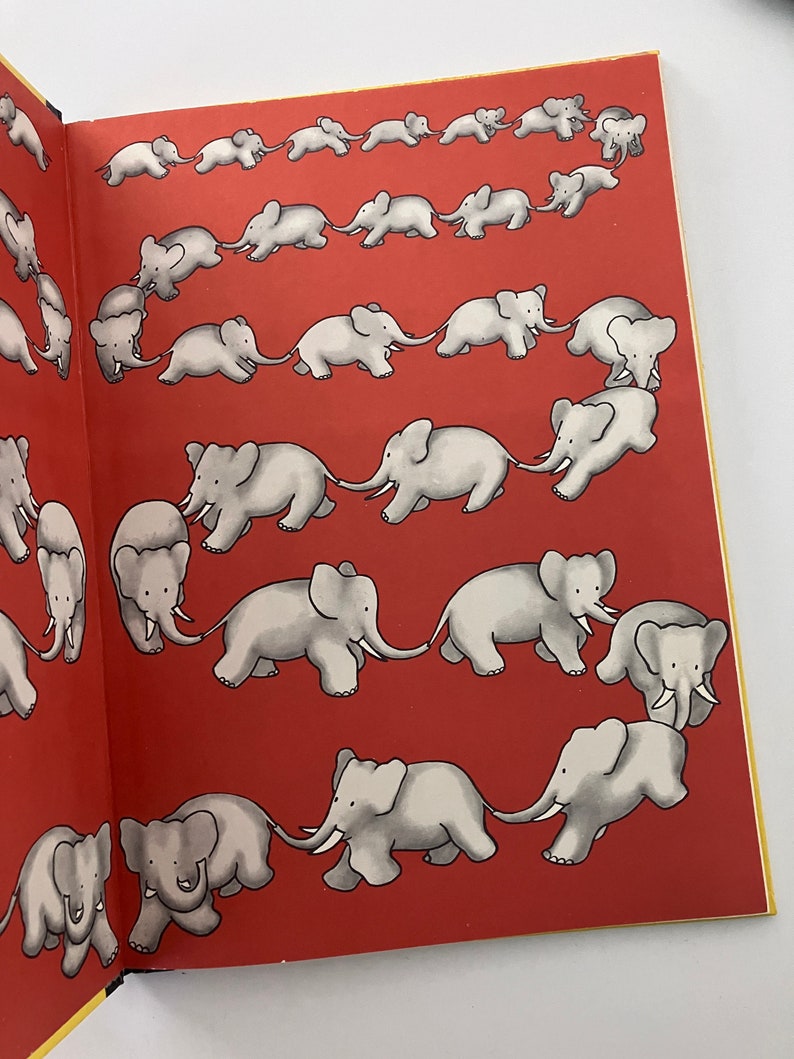 May include: A red book with a yellow border, featuring a series of illustrations of elephants walking in a line, with each elephant holding the tail of the elephant in front of it. The elephants are all facing the same direction and are drawn in a simple, cartoon style.