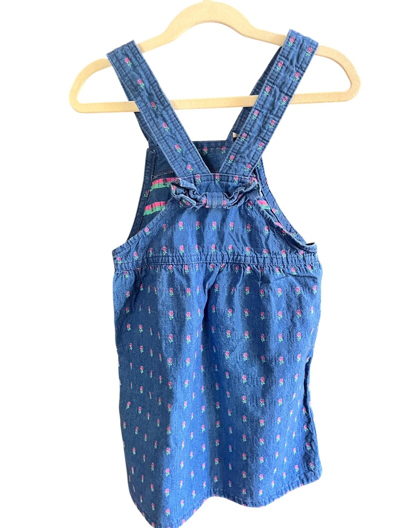 Op de afbeelding: Blauwe denim overall jurk met roze bloemenpatroon en ruches. De bandjes en de achterkant hebben een bloemenmotief. De jurk hangt aan een houten hanger.
