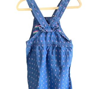 Op de afbeelding: Blauwe denim overall jurk met roze bloemenpatroon. De jurk heeft verstelbare bandjes en een gerimpeld detail aan de achterkant. Het kledingstuk hangt aan een houten hanger.