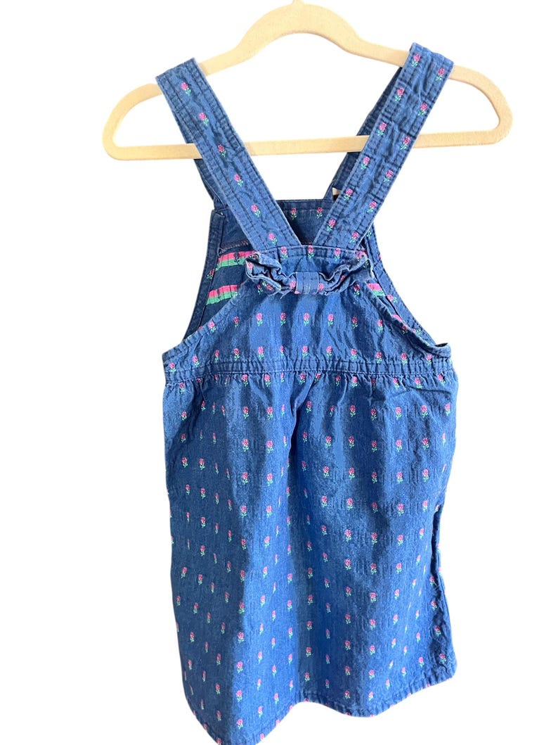 Op de afbeelding: Blauwe denim overall jurk met roze bloemenpatroon. De jurk heeft verstelbare bandjes en een gerimpeld detail aan de achterkant. Het kledingstuk hangt aan een houten hanger.