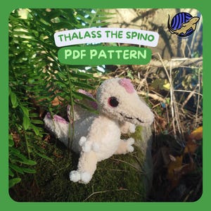 May include: A crocheted dinosaur toy with a cream-colored body, pink accents, and large red eyes. The text "THALASS THE SPINO PDF PATTERN" is displayed above the dinosaur. The toy is set against a backdrop of green foliage.