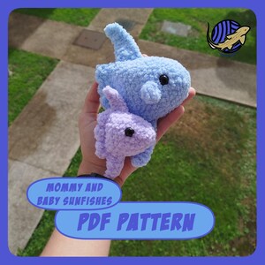 May include: A light blue and lavender crocheted sunfish, with black eyes, held in a hand. The larger sunfish is light blue, and the smaller one is lavender. Text on the image reads "MOMMY AND BABY SUNFISHES PDF PATTERN".