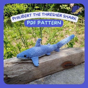 May include: A blue crocheted shark toy with the text "PHILLIBERT THE THRESHER SHARK PDF PATTERN". The shark is lying on a weathered wooden beam, with green foliage in the background. The shark has a long tail and fins.