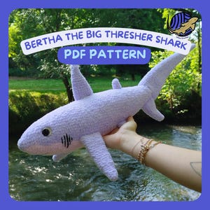May include: A light purple crocheted thresher shark plush toy with black embroidered gills and dark eyes. The text BERTHA THE BIG THRESHER SHARK PDF PATTERN is displayed in blue letters on a white curved banner above the shark.