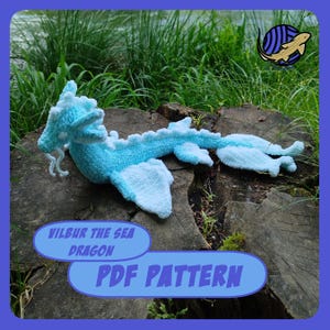 Op de afbeelding: Een gehaakte zeedraak in lichtblauw en wit, liggend op een boomstronk. De draak heeft een lang lichaam, vleugels en een gedetailleerde kop. De afbeelding bevat de tekst "VILBUR THE SEA DRAGON PDF PATTERN".
