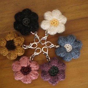 May include: A collection of six crocheted flower charms in various colors, including black, white, yellow, blue, pink, and purple. Each flower has a silver-colored lobster clasp attached, perfect for use as keychains or bag charms. The flowers are arranged on a wooden surface.