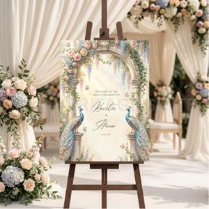 May include: A wooden easel displays a wedding welcome sign. The sign features an archway design with floral accents, two peacocks, and the names "Kavita & Arun." The sign is surrounded by floral arrangements and draped fabric.