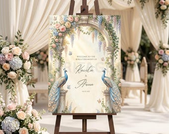 May include: A wooden easel displays a wedding welcome sign. The sign features an archway design with floral accents, two peacocks, and the names "Kavita & Arun." The sign is surrounded by floral arrangements and draped fabric.