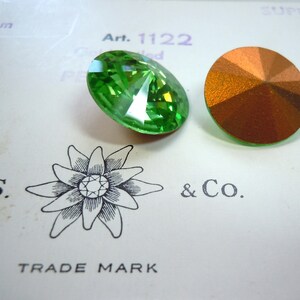 May include: Two round, faceted glass rhinestones. One is green and the other is copper. The rhinestones are on a white card with the text "Art. 1122" and "D.S. & Co. TRADE MARK" printed on it.