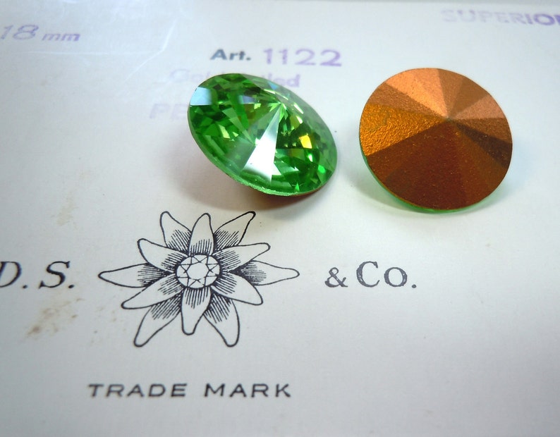May include: Two round, faceted glass rhinestones. One is green and the other is copper. The rhinestones are on a white card with the text "Art. 1122" and "D.S. & Co. TRADE MARK" printed on it.