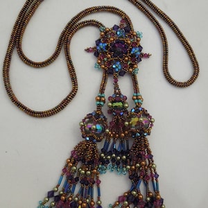 Puede incluir: Un collar de cuentas con un gran colgante en forma de estrella multicolor y dos borlas colgantes. El collar está hecho con cuentas marrones, moradas, azules y doradas.