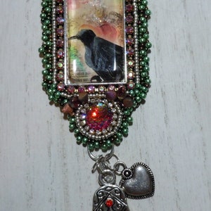 May include: A silver necklace with a rectangular pendant featuring a black crow on a pink and green background. The pendant is surrounded by green and brown beads. The necklace also has a silver charm in the shape of a hamsa hand with red and green stones and a silver heart charm.