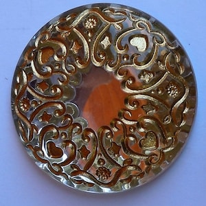 May include: A clear, round decorative object with an intricate gold filigree design. The filigree features swirling patterns, hearts, and star shapes. The center of the object has a reflective, copper-colored appearance. The object is likely a decorative element or embellishment.