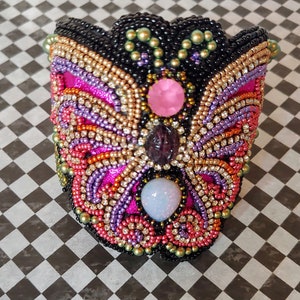 May include: A beaded butterfly brooch with pink, purple, orange, and gold accents. The butterfly has three large gemstones in the center.