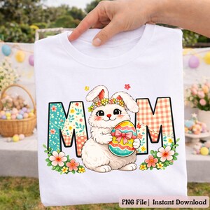 May include: White t-shirt featuring a cartoon bunny holding a decorated Easter egg. The word "MOM" is displayed with floral and plaid patterns. The bunny wears a flower crown, and the design is surrounded by flowers.
