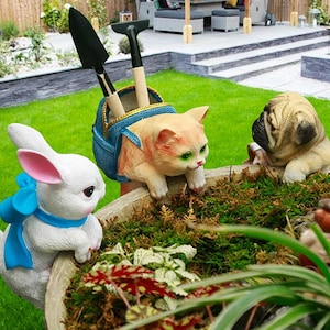 Könnte beinhalten: Ein Gartenpflanzer mit drei Tierfiguren: ein weißes Kaninchen mit blauer Schleife, eine hellbraune Katze mit grünen Augen und eine Möpse. Die Katze hält eine blaue Tasche mit Gartengeräten. Der Pflanzer ist mit Grünzeug gefüllt.
