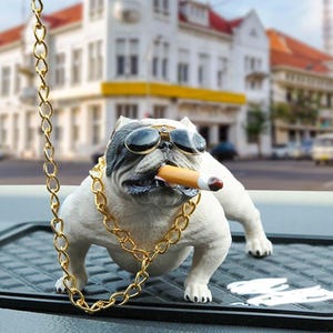 Könnte beinhalten: Eine weiß-graue Bulldoggenfigur mit Sonnenbrille und goldener Kette, mit einer brennenden Zigarre im Maul. Die Figur steht auf einer schwarzen, strukturierten Oberfläche, mit einer goldenen Kette, die von oben hängt.